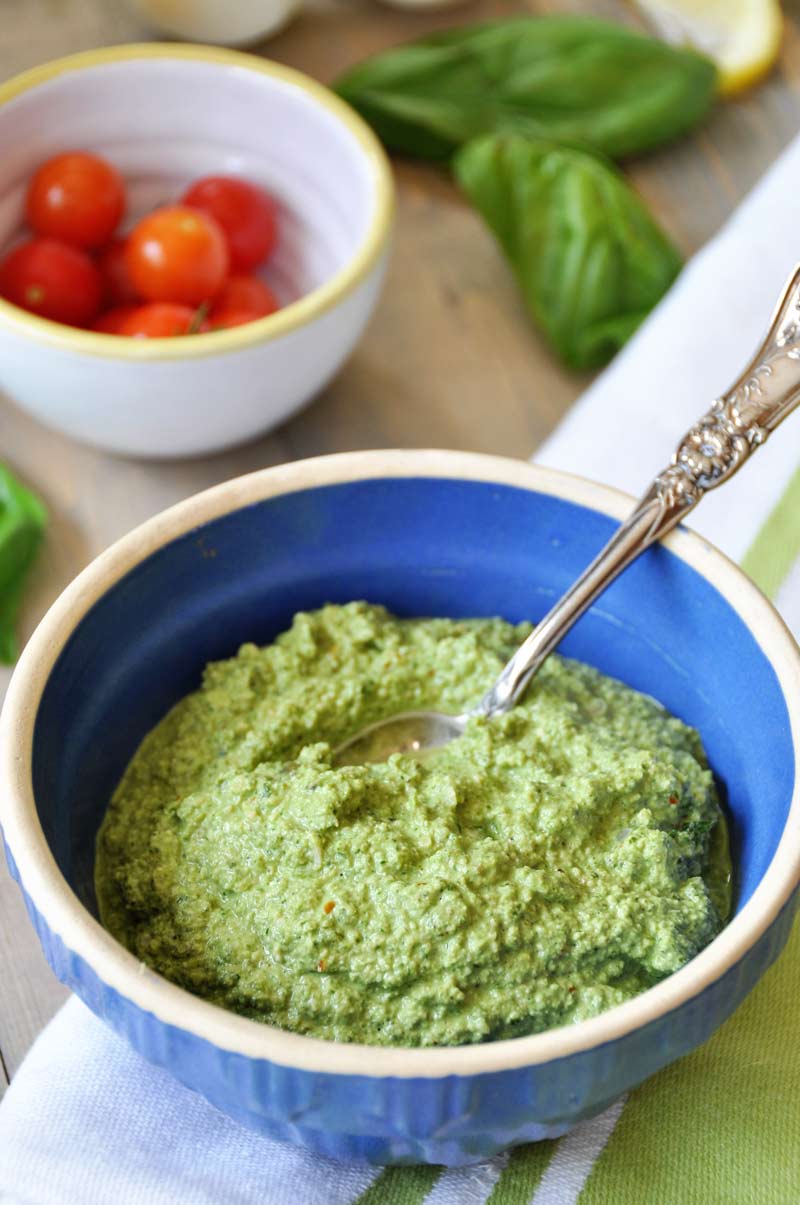 A blue bowl with green sauce and a silver spoon in it, and a white bowl with tomatoes, and a bunch of basil in the background.