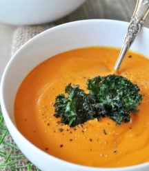 Carrot soup topped with kale chips in a white bowl with a silver spoon in the bowl. Sprigs of rosemary and another bowl of soup are next to the bowl of soup.