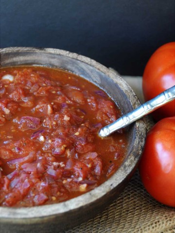 A wooden bowl filled with red tomato spaghetti sauce with a silver spoon in the bowl and tomatoes to the right of the bowl.