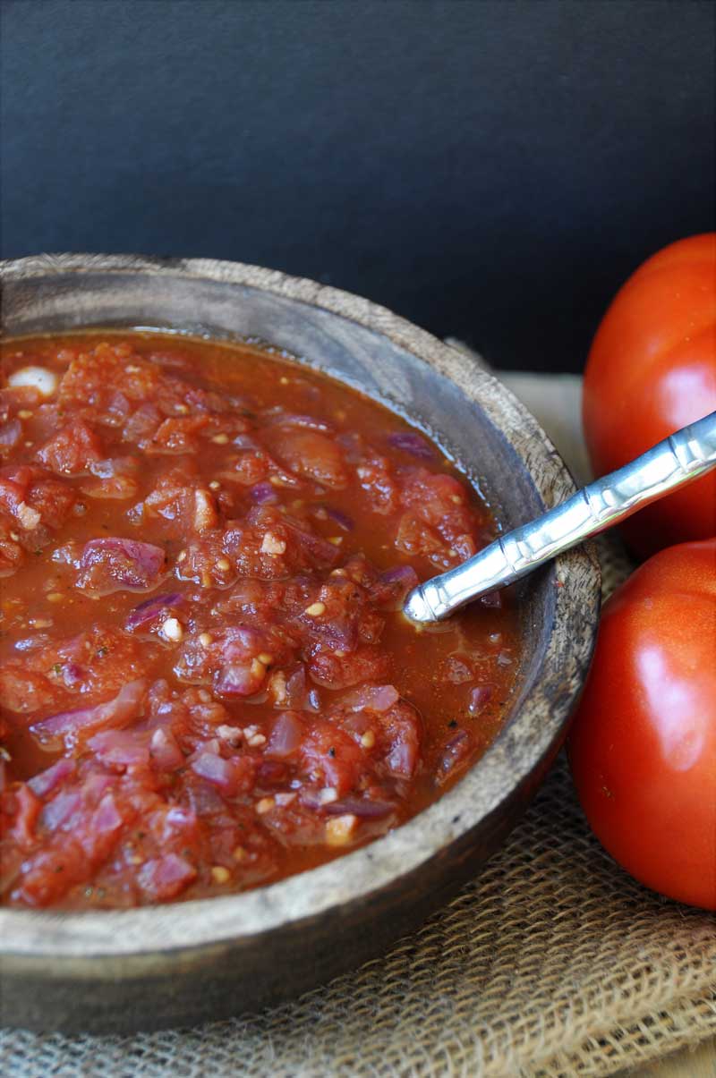 A wooden bowl filled with red tomato spaghetti sauce with a silver spoon in the bowl and tomatoes to the right of the bowl.