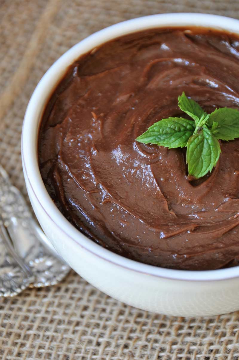 Silky smooth chocolate pudding in a white bowl with a sprig of mint on top on a burlap cloth with two silver spoons next to it.