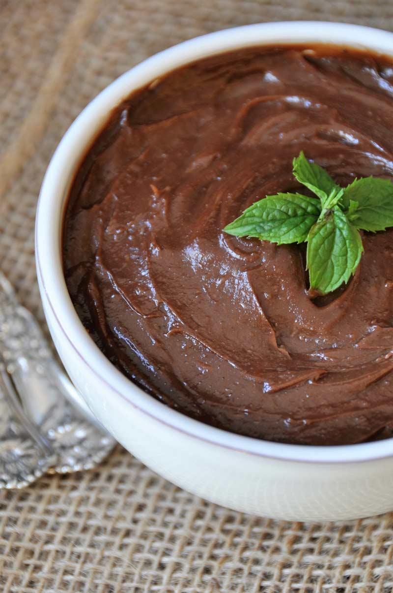 Silky smooth chocolate pudding in a white bowl with a sprig of mint on top on a burlap cloth with two silver spoons next to it. 