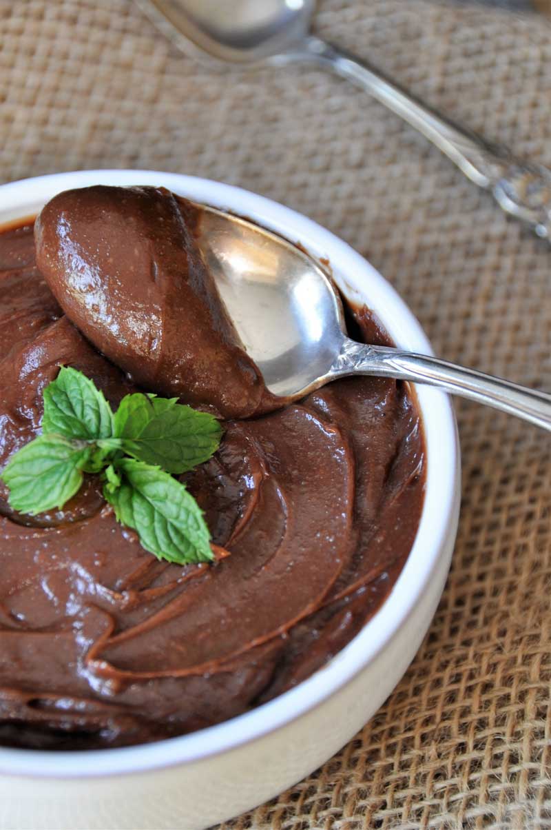 Silky smooth chocolate pudding in a white bowl with a sprig of mint and a silver spoon full of pudding on the edge of the bowl.