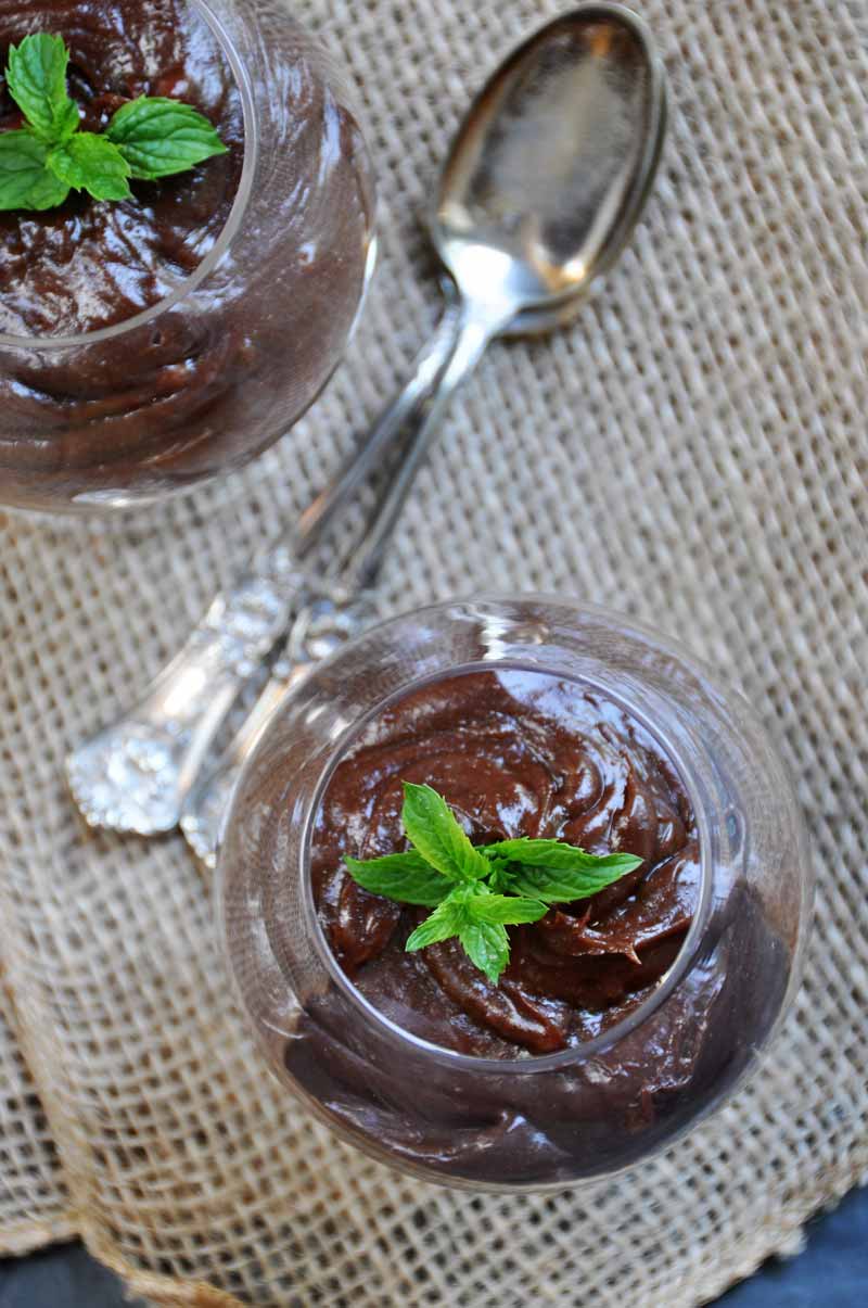 Two glass cups of silky smooth chocolate pudding with sprigs of mint on top, on a burlap cloth with two silver spoons between the cups.