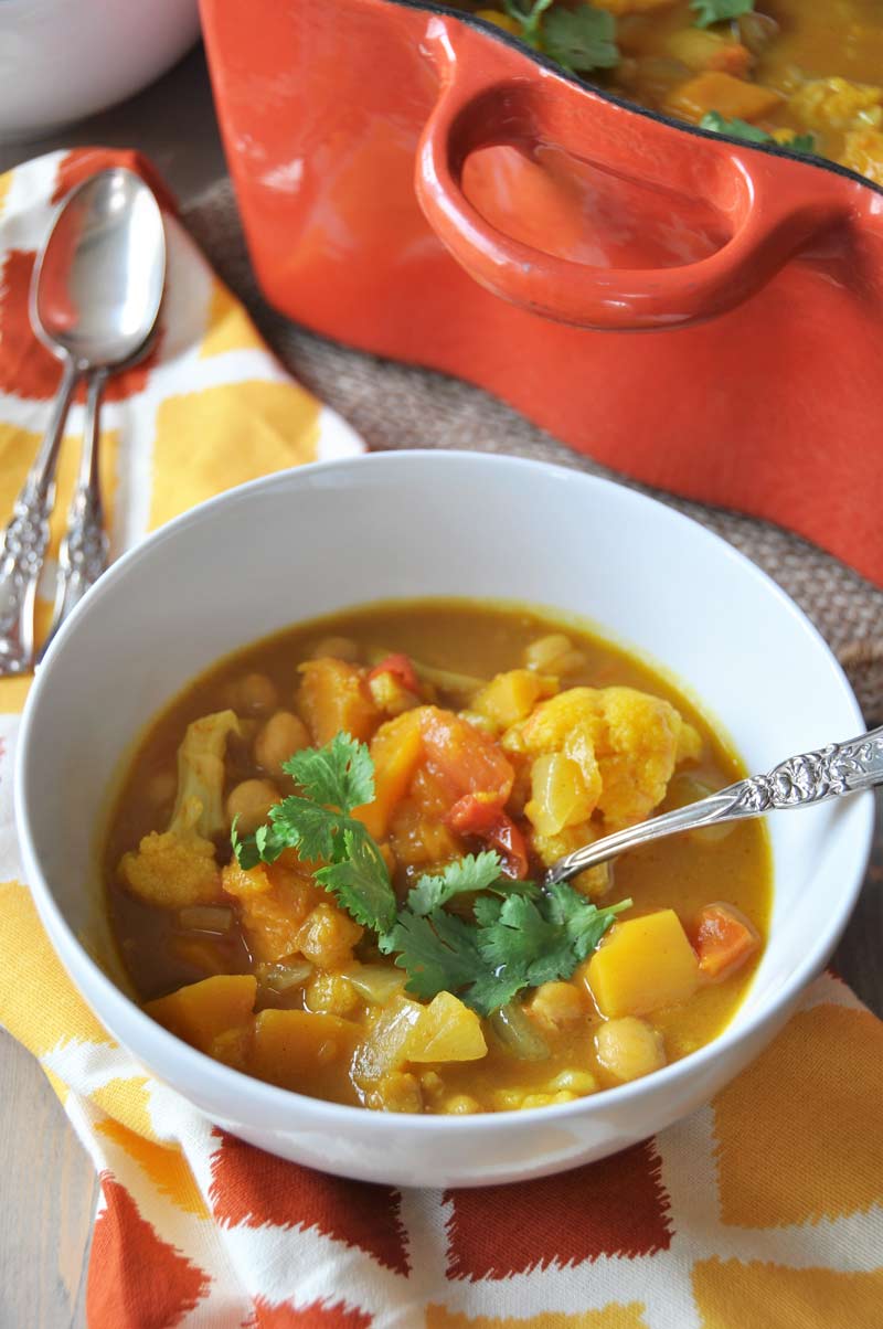 Curried pumpkin and butternut squash stew in a white bowl with a sprig of cilantro on top and a silver spoon in the bowl, sitting next to an orange Dutch oven filled with stew. 