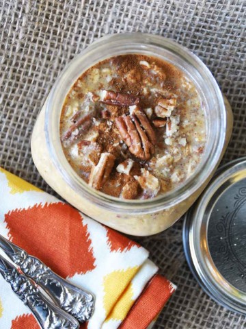 pumpkin pie overnight oats in a mason jar with pecans on top and an orange and gold napkin and silver spoons next to it.