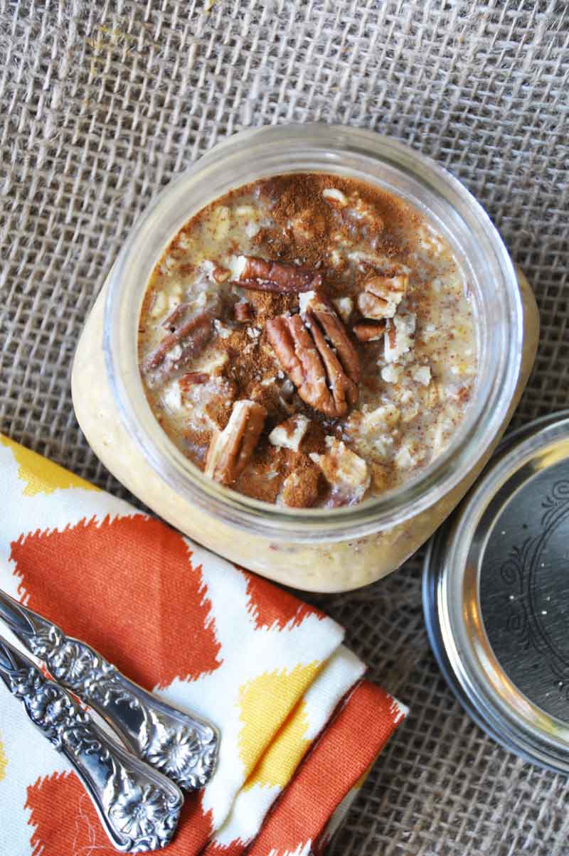 pumpkin pie overnight oats in a mason jar with pecans on top and an orange and gold napkin and silver spoons next to it. 