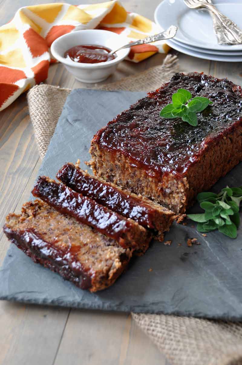 Sliced Vegan meatloaf on a slate tray with fresh herbs next to a yellow and orange napkin and a white dish of extra sauce and plates and forks