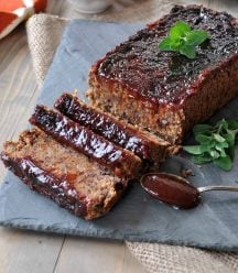 A vegan meatloaf with BBQ Sauce on a slate platter with a spoon of sauce and fresh herbs