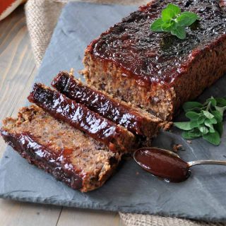 A vegan meatloaf with BBQ Sauce on a slate platter with a spoon of sauce and fresh herbs