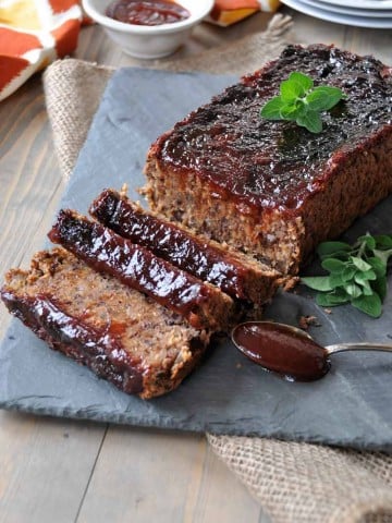 A bean meatloaf with bbq sauce and a sprig of oregano on top of it with a silver spoon dripping bbq sauce next to the loaf