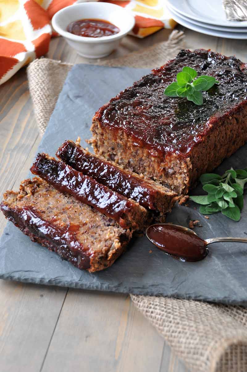 Sliced Vegan meatloaf on a slate platter with a spoon of sauce next to it and fresh sprigs of oregano and a dish of BBQ sauce and white plates in the background