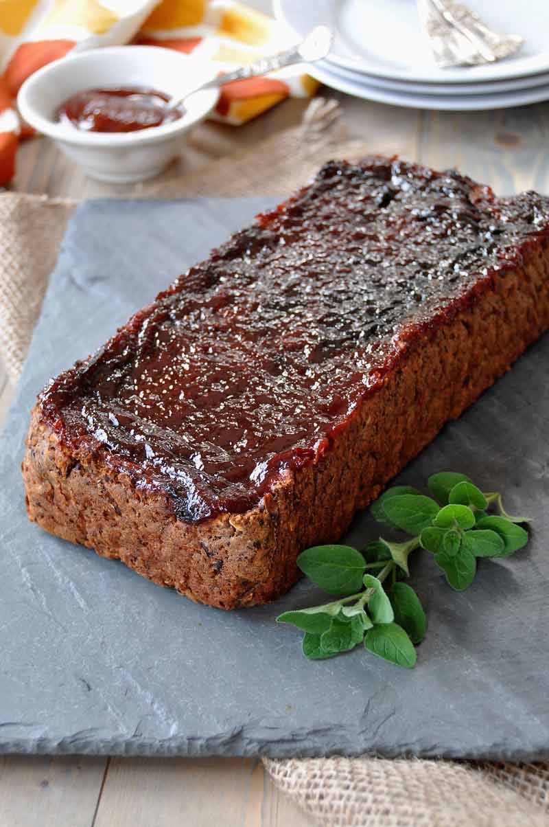 Vegan meatloaf on slate plate with a sprig of fresh oregano and a white dish of BBQ sauce and white plates and silver forks behind it.