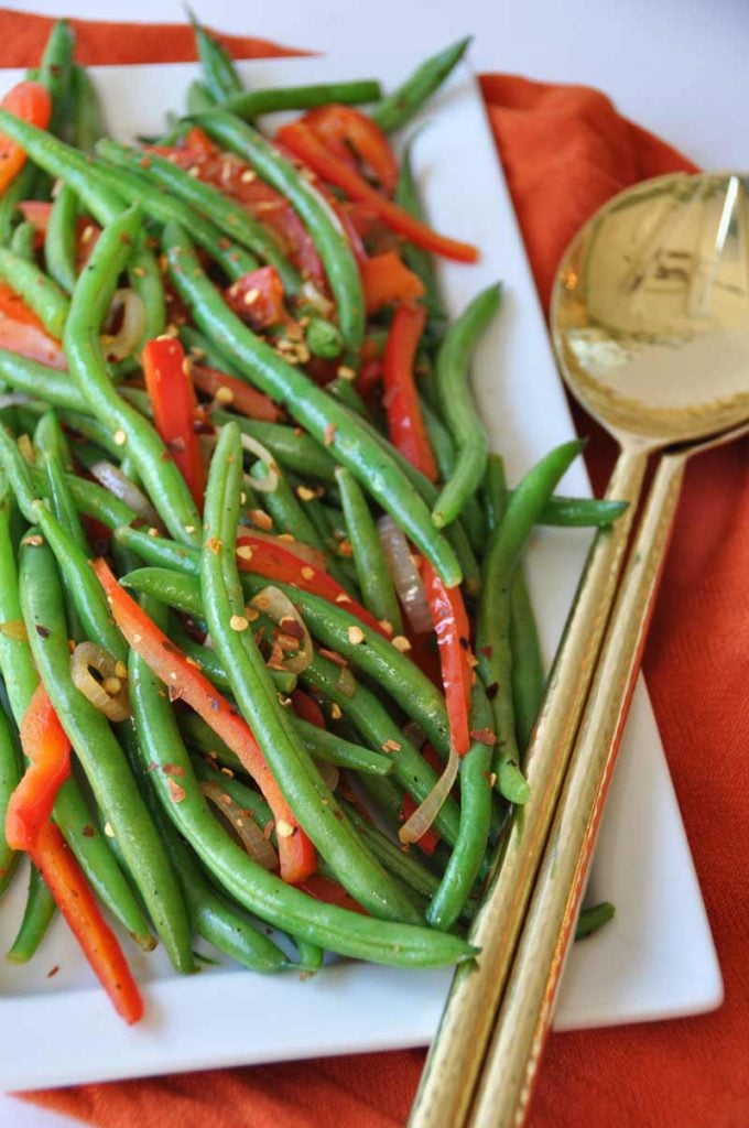 Spicy flash fried green beans with red peppers and pepper flakes on a white platter with gold utensils resting on the edge of the platter on an orange cloth