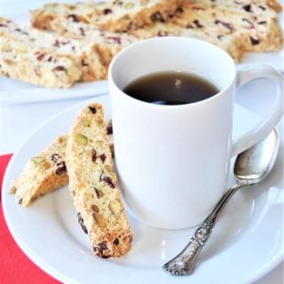 A white mug with coffee on a white saucer with a silver spoon and 2 biscotti cookies on the saucer, and a platter of the biscotti in the background.