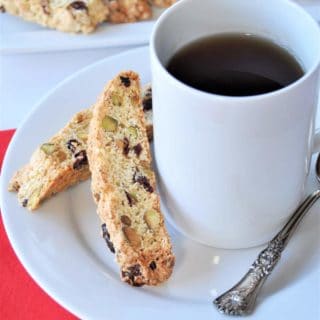 Cranberry biscotti on a white plate next to a white ceramic cup of coffee with a silver spoon resting on the edge of the plate and a red napkin next to the plate and a white plate of cookies in the background