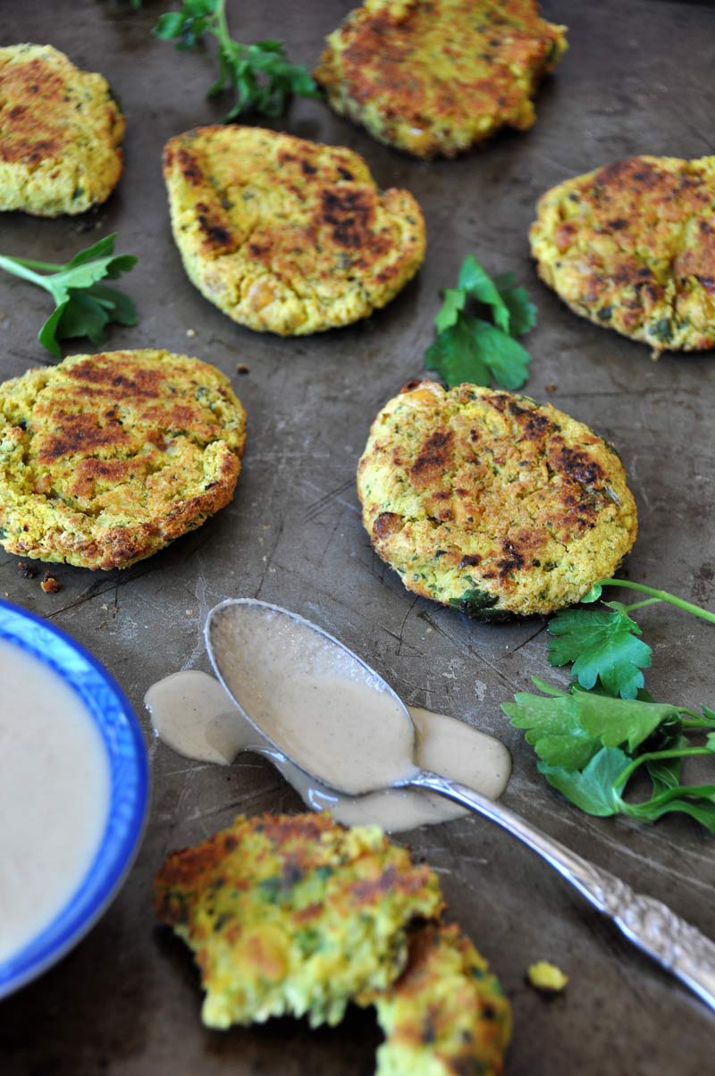 Crispy oven fried falafel spread on a baking sheet with cilantro sprigs and a blue bowl with tahini and a spoon next to it with tahini dripping off of it