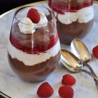 Two glasses of chocolate parfaits with whip cream and raspberries on a marble tray with two silver spoons and raspberries on the tray.