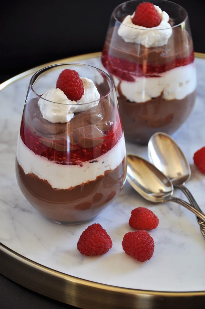 Two raspberry parfaits in stemless wine glasses with two silver spoons and raspberries on a marble tray.