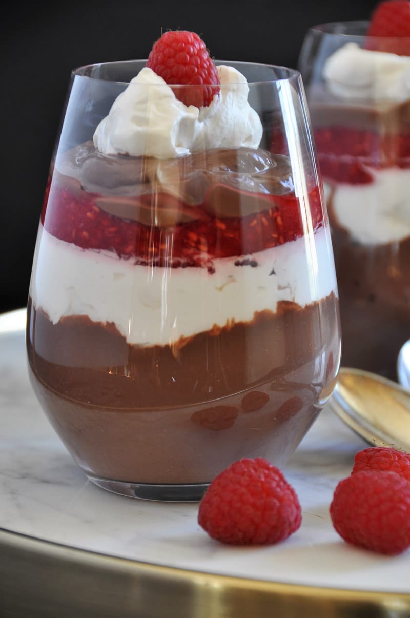 A glass with a chocolate raspberry parfait with another one in the background and raspberries on a marble tray