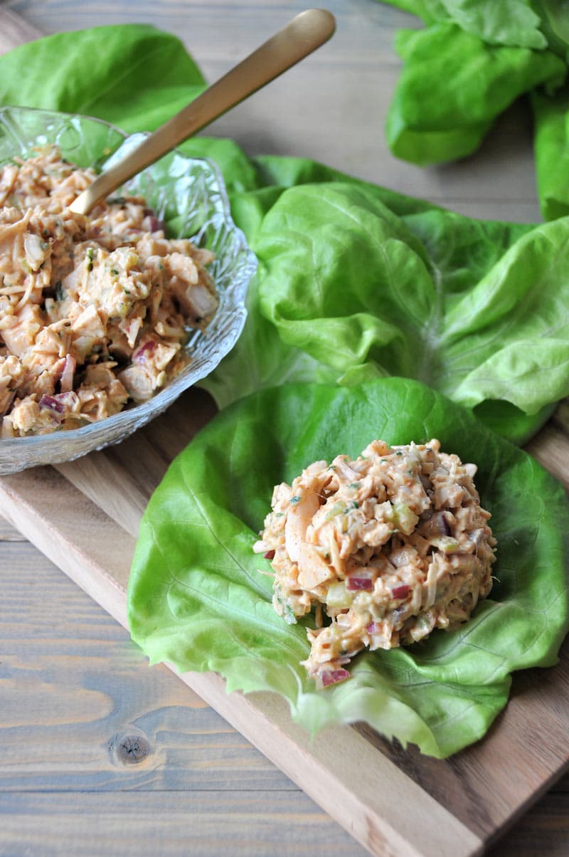 Jackfruit tuna-less salad piled on a lettuce leaf on a wood board with several other leaves next to it and a clear glass dish with the salad in it and a gold spoon