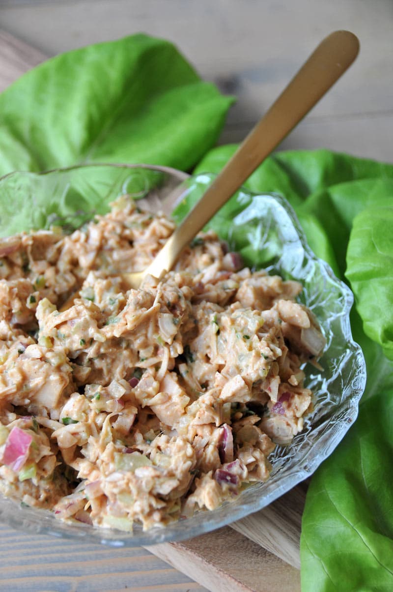 A clear glass bowl with jackfruit tuna-less salad and a gold spoon sticking out of it with lettuce leaves surrounding the bowl.