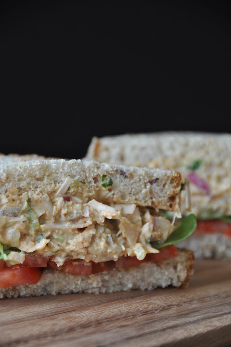 A jackfruit tuna-less salad sandwich cut in half with whole grain bread, lettuce and tomato on a wood board