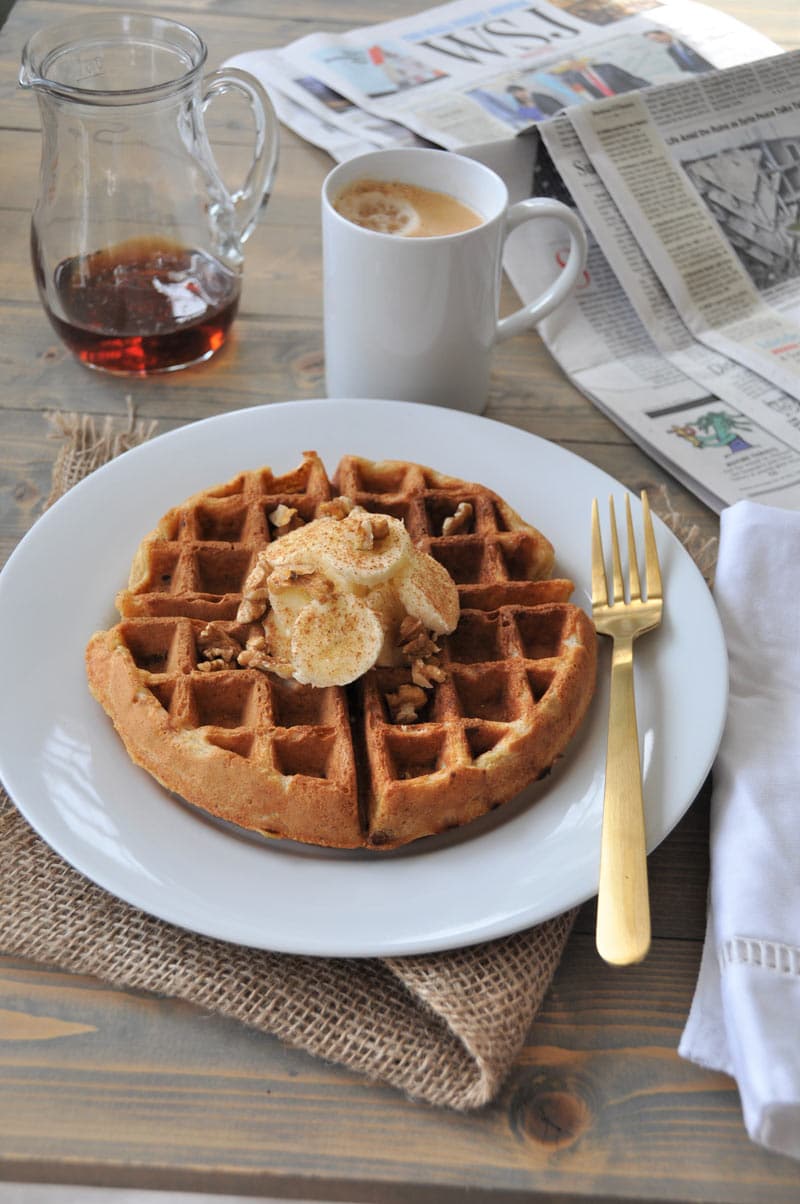 A Belgian waffle with sliced bananas and nuts on a white plate with a gold fork on the side, and a pitcher with maple syrup and a white mug of coffee in the background.
