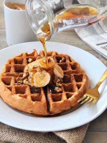 A clear glass pitcher with maple syrup being poured on a Belgian waffle with sliced bananas and nuts.