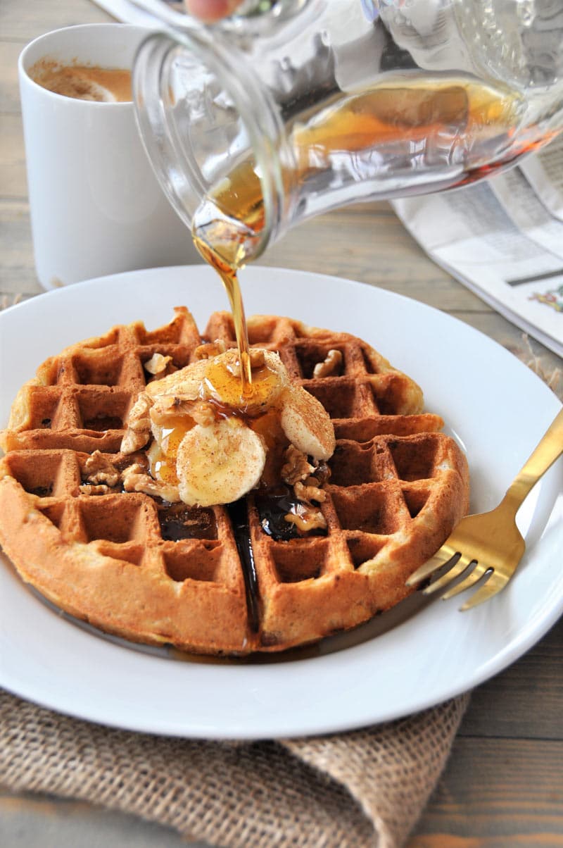 A clear glass pitcher with maple syrup being poured on a Belgian waffle with sliced bananas and nuts.