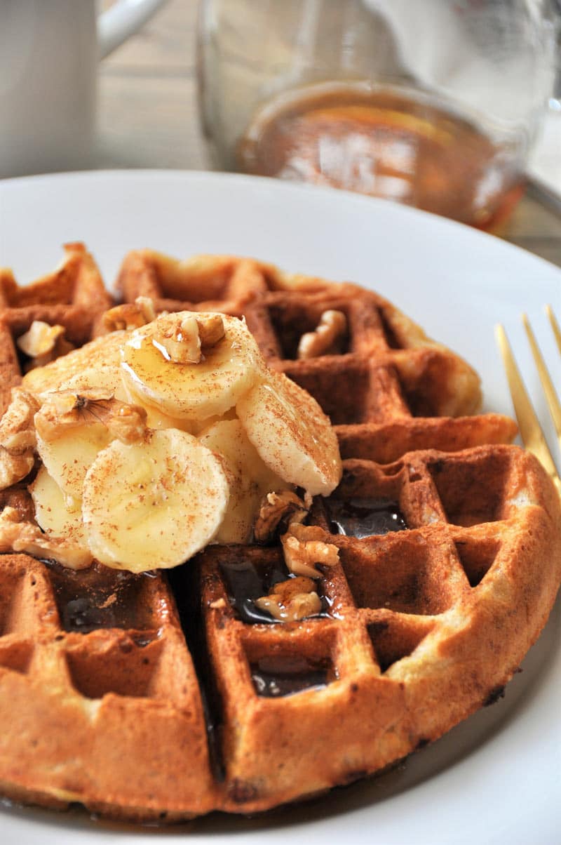 A picture of a Belgian waffle on a white plate with sliced bananas, nuts, and maple syrup.