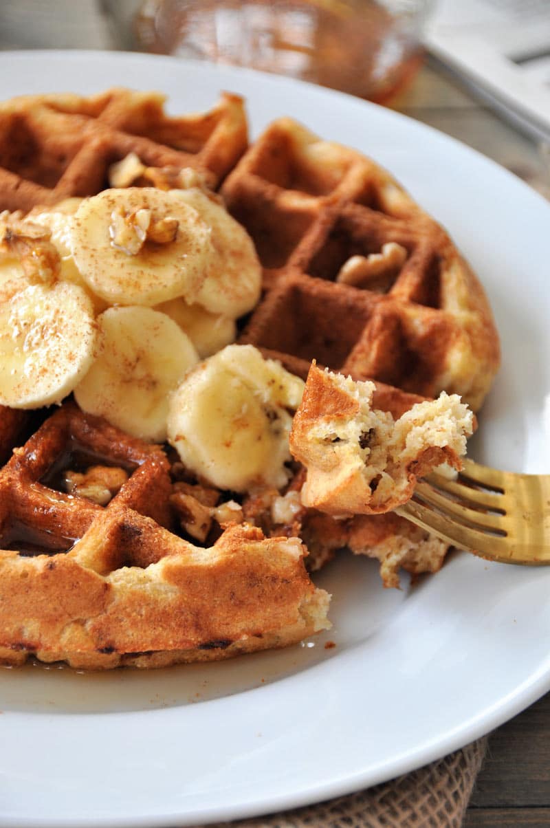 A gold fork with a piece of Belgian waffle and the rest of the whole waffle topped with banana slices and nuts on a white plate.