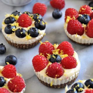 Cheesecake bites with blueberries, and raspberries scattered on a marble surface with a white scalloped bowl of blueberries and raspberries in the background