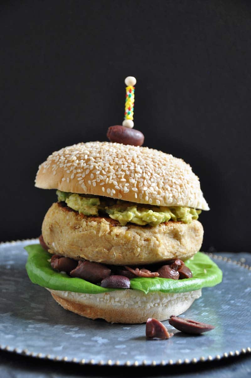 Hummus burger on a silver plate with an olive stuck on the top of a sesame seed bun with a toothpick