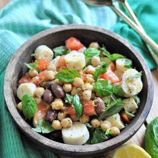 Chickpea salad with vegetables in a wood bowl with a green napkin around it and gold serving utensils and lemon wedges on the side