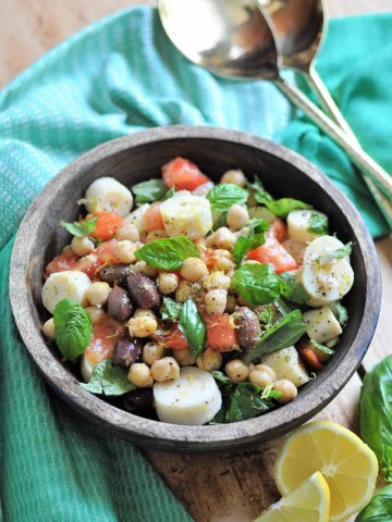 Chickpea salad with vegetables in a wood bowl with a green napkin around it and gold serving utensils and lemon wedges on the side