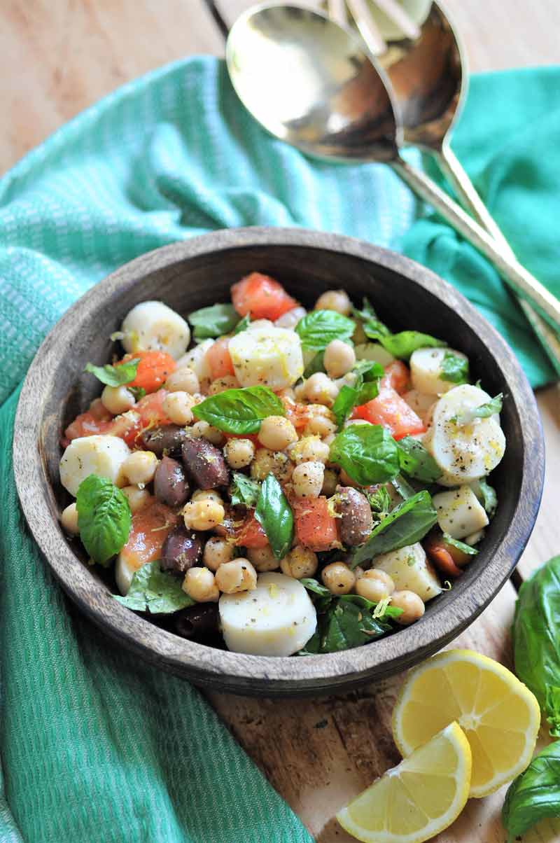 Chickpea salad with vegetables in a wood bowl with a green napkin around it and gold serving utensils and lemon wedges on the side