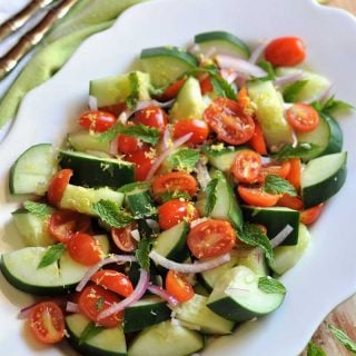 Cucumber, Tomato & Mint Salad on a scalloped edged white platter with a green and white napkin with silver serving utensils next to it