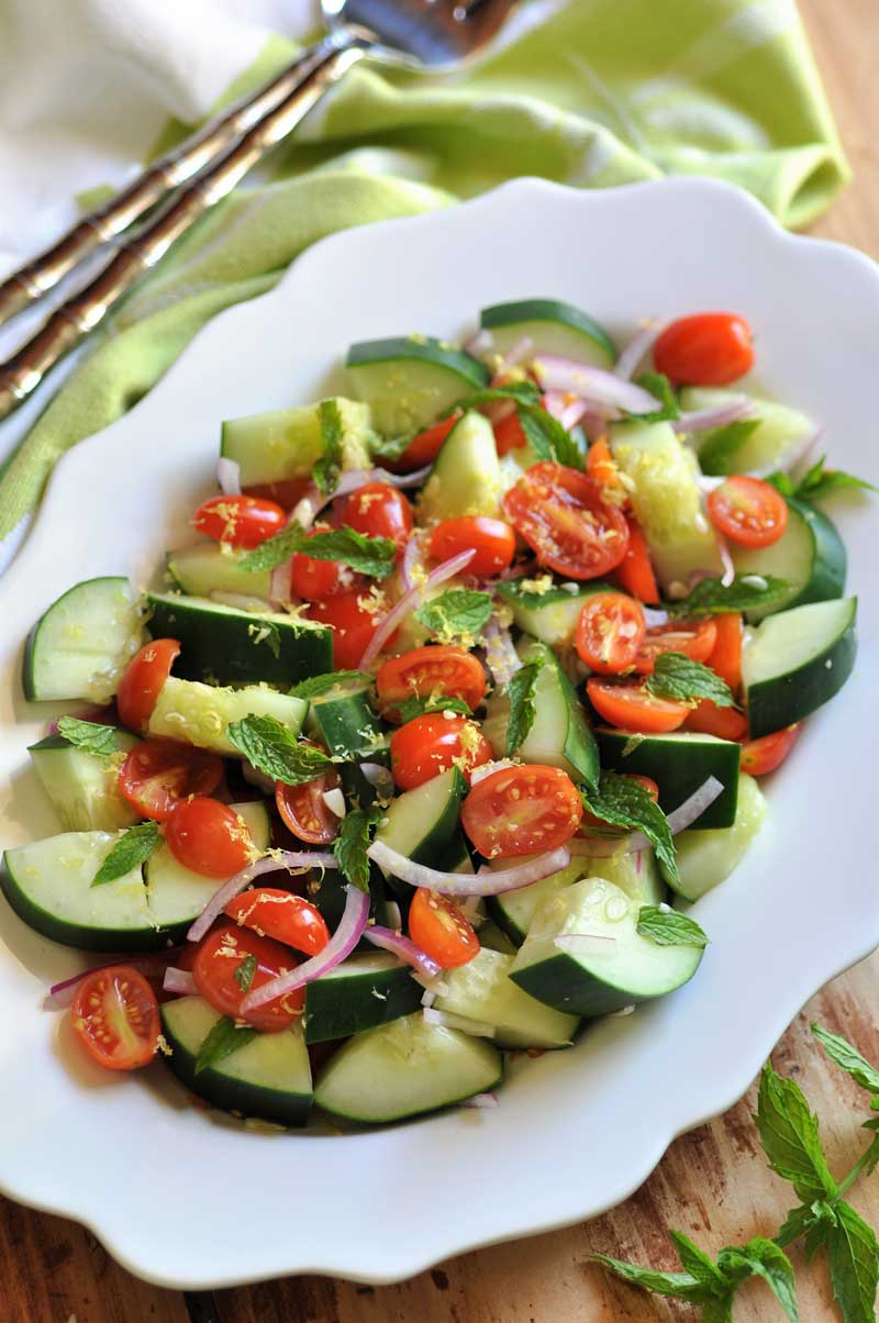 Cucumber, Tomato & Mint Salad on a scalloped edged white platter with a green and white napkin with silver serving utensils next to it