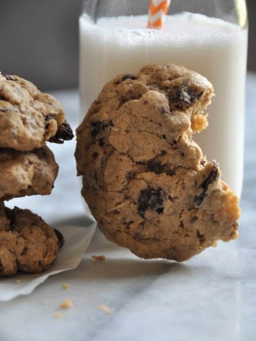 An oatmeal raisin cookie with a bite out of it, leaning on a glass of milk.