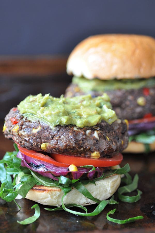 An open face black bean veggie taco burger with lettuce and tomatoes and guacamole spread on top of the burger, and another burger with the bun on top behind it.