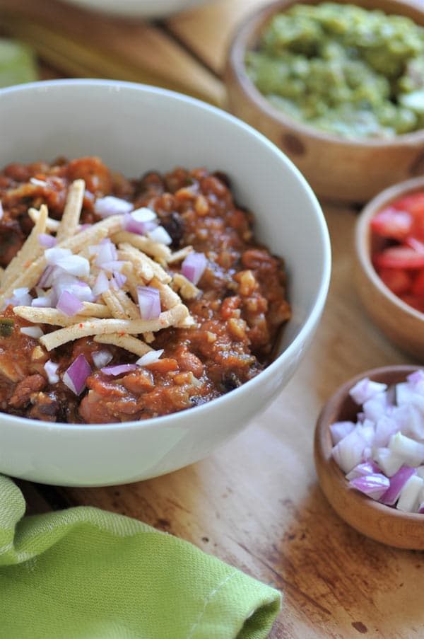 Hearty veggie chili in a white bowl with vegan cheese shreds and onion on top and three wood bowls filled with onion, tomato, and guacamole next to it