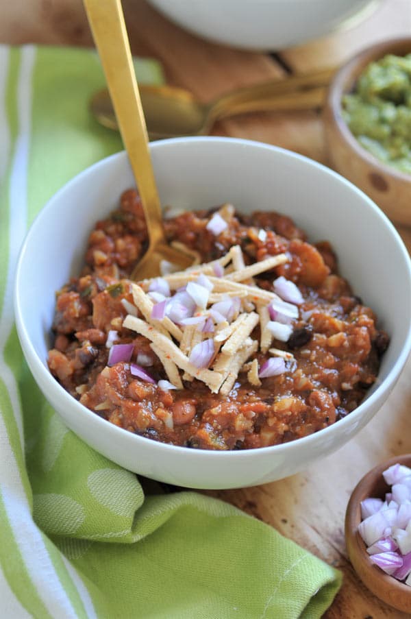 Vegan chili in a white bowl with shredded vegan cheese and diced onion on top with a gold spoon in the bowl and a green napkin on the side