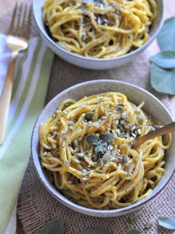 Pumpkin sage cream sauce smothered over spaghetti in a white bowl, sprinkled with pepitas and sage leaves.