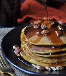 A stack of five vegan pumpkin pancakes on a black plate, sprinkled with pecans and a pat of butter. Syrup is being poured over them.