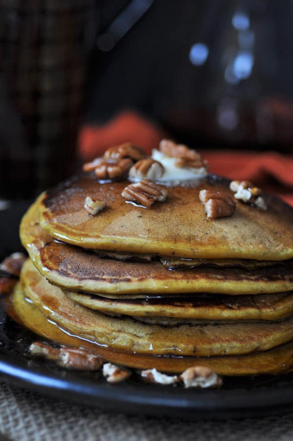 Five vegan pumpkin pancakes with a pat of vegan butter melting on top, with pecans and maple syrup
