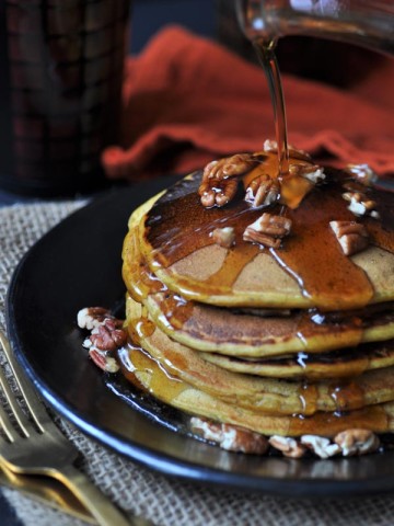A stack of five vegan pumpkin pancakes on a black plate, sprinkled with pecans and a pat of butter. Syrup is being poured over them.