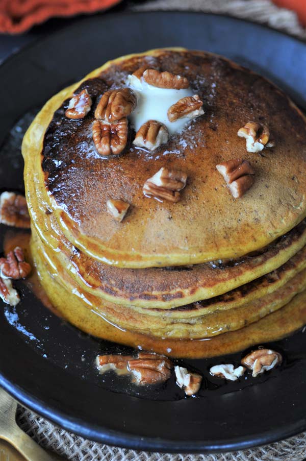 An overhead view of a stack of vegan pumpkin pancakes with maple syrup, pecans, and melting butter