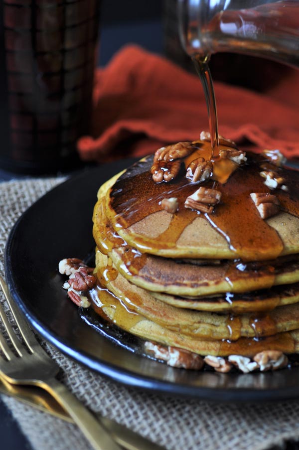 A stack of five vegan pumpkin pancakes on a black plate, sprinkled with pecans and a pat of butter. Syrup is being poured over them.