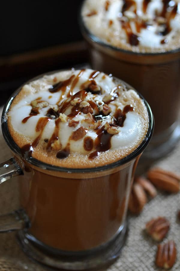 Homemade Vegan Maple Pecan Lattes with whipped topping and a drizzle of maple syrup and pecans scattered on a burlap cloth